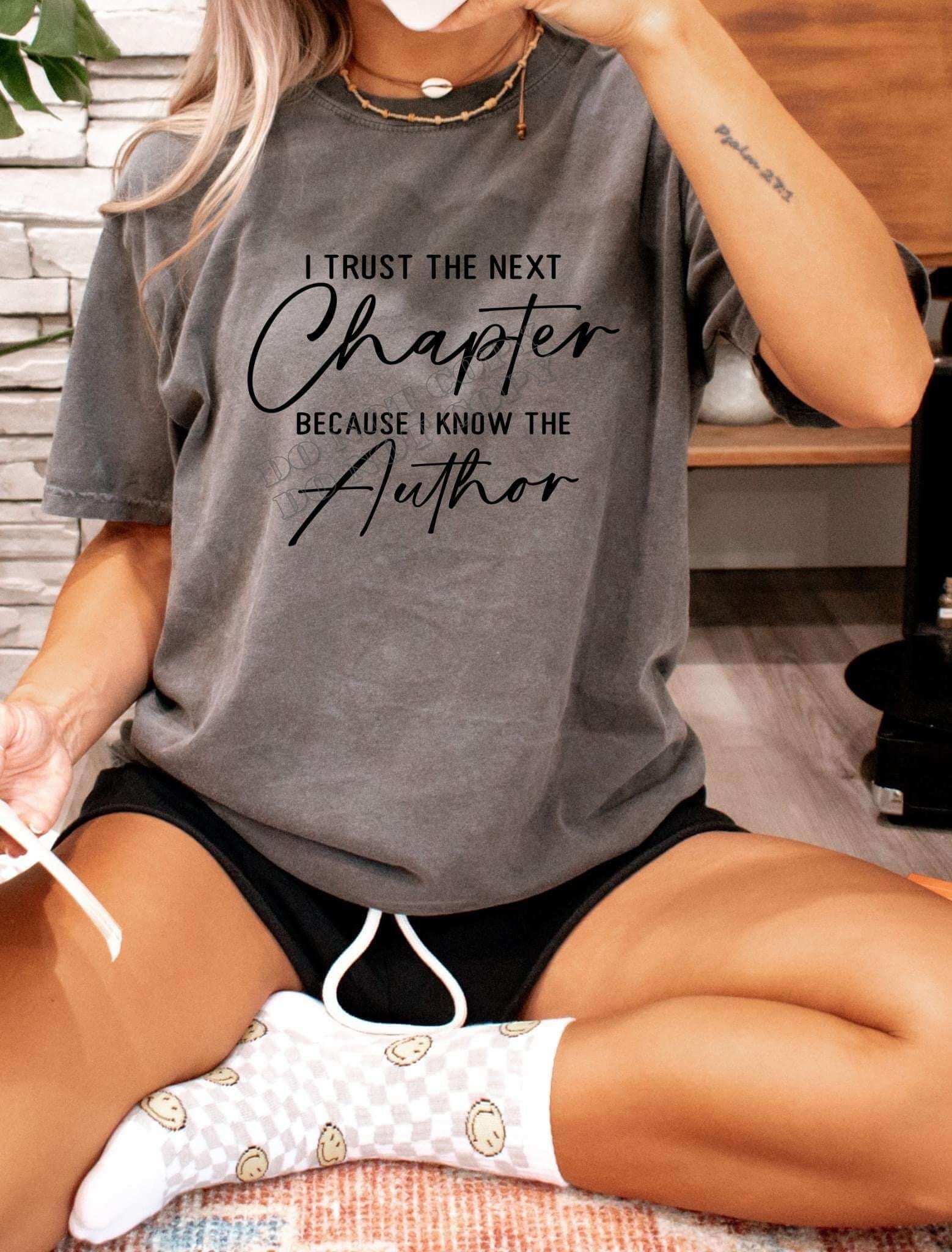 Woman wearing a charcoal gray Christian T-shirt with the quote 'I Trust the Next Chapter Because I Know the Author' while sitting on the floor in cozy socks and shorts