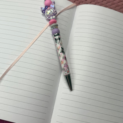 Purple pastel kitty beaded pen with black ink resting on an open lined journal, styled for girly desk décor.