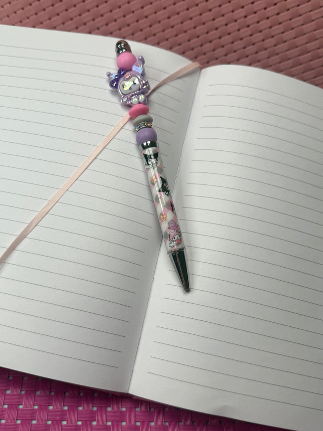 Purple pastel kitty beaded pen with black ink resting on an open lined journal, styled for girly desk décor.