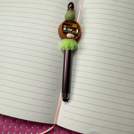 Stylus pen in maroon with sage green silicone bead and rhinestone spacer with center focal of round bookshelf with sleeping cat, books and potted plant. 
