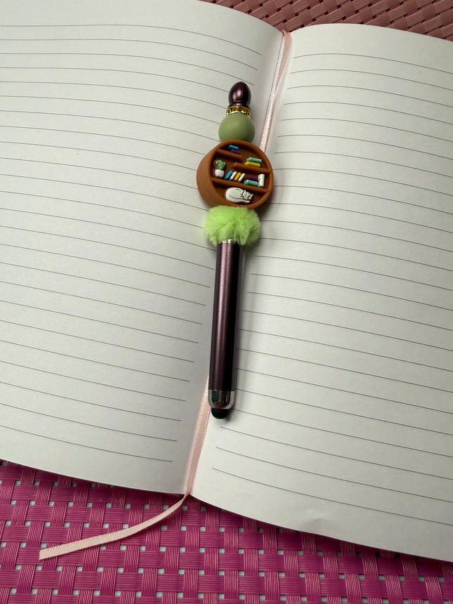 Stylus pen in maroon with sage green silicone bead and rhinestone spacer with center focal of round bookshelf with sleeping cat, books and potted plant. 