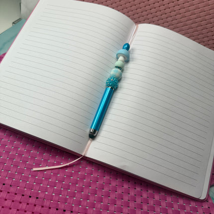 Metallic Bright blue stylus pen with blue glittery rhinestone with blue and white plaid silicone bead and solid light blue bead with a light blue white spotted mushroom laying an open journal book. 