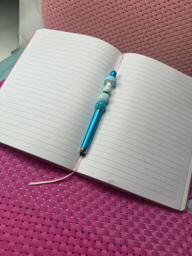 Metallic Bright blue stylus pen with blue glittery rhinestone with blue and white plaid silicone bead and solid light blue bead with a light blue white spotted mushroom laying an open journal book. 