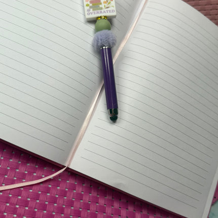 Purple Stylus pen laying on open notebook. Features a silicone focal that says, “Books because reality is over rated. Has a stack of books with flowers around statement with sage green round silicone bead and rhinestone spacer