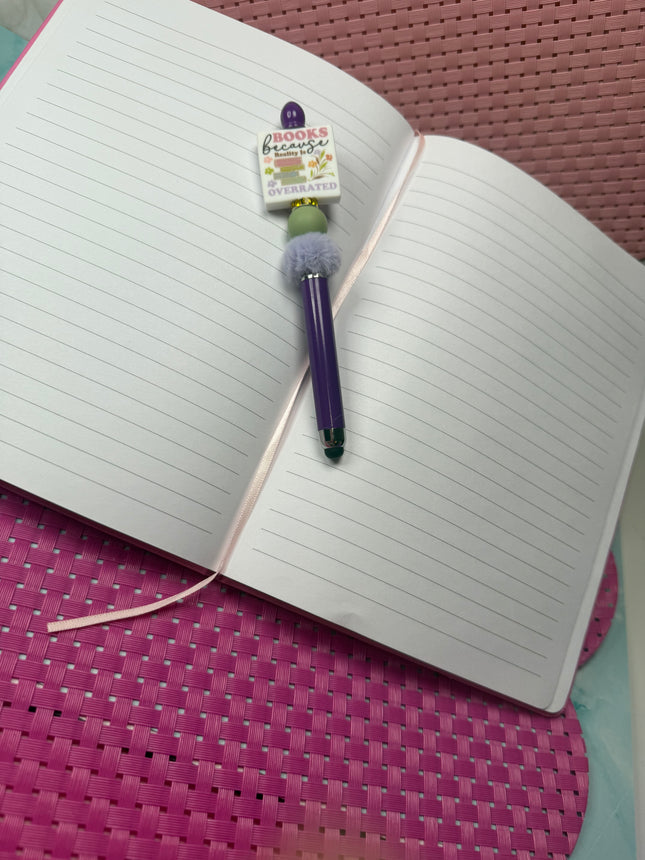 Purple Stylus pen laying on open notebook. Features a silicone focal that says, “Books because reality is over rated. Has a stack of books with flowers around statement with sage green round silicone bead and rhinestone spacer