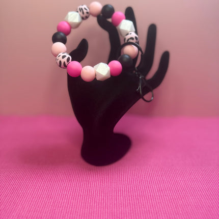 A beaded stretch bracelet with pink, white, and black beads, displayed on a black hand-shaped jewelry stand against a pink background.