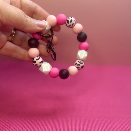 A hand holding a beaded keychain bracelet with pink, black, and leopard-print beads against a pink background.