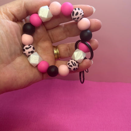 A hand holding a beaded stretch bracelet with pink, light pink, black, and leopard-print beads against a pink background.