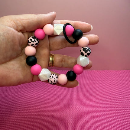 A hand holding a beaded stretch bracelet with pink, black, and white leopard-print beads against a pink background.