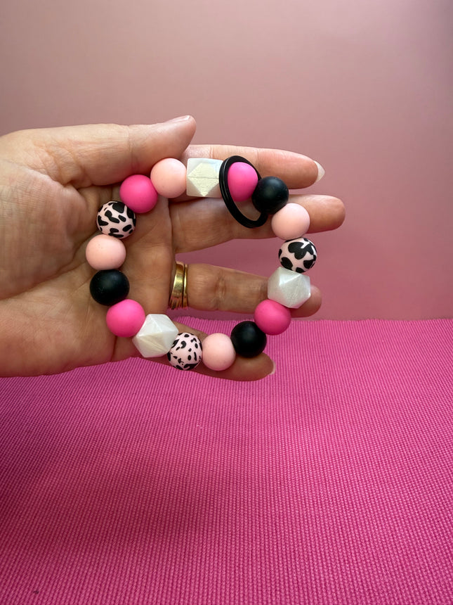 A hand holding a beaded stretch bracelet with pink, black, and white leopard-print beads against a pink background.