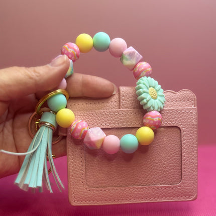 A hand holds a pastel beaded card holder keychain with a tassel and a small flower charm in front of a pink textured background.