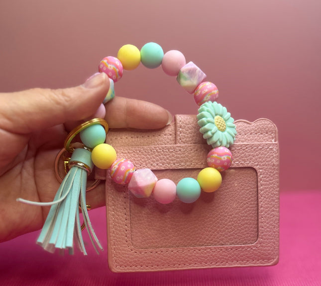 A hand holds a pastel beaded card holder keychain with a tassel and a small flower charm in front of a pink textured background.