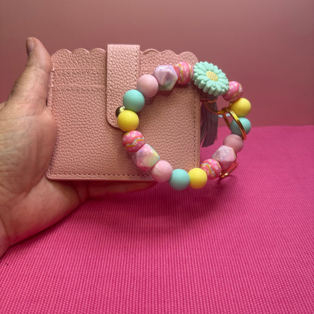 A hand holds a pink textured card holder wallet with a pastel beaded bracelet draped over it. The bracelet features pastel pink, mint green, yellow, and marbled beads, along with a small daisy charm and a feather accent, set against a pink background.