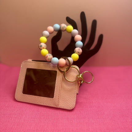 A pink faux-leather card holder keychain with a colorful beaded wrist strap and gold key rings, displayed on a pink surface with a black hand-shaped backdrop against a peach background.