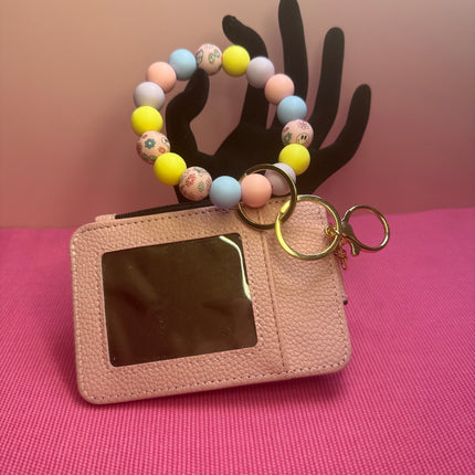 A pink textured leather card holder keychain with a clear ID window, gold key rings, and a colorful beaded wrist strap, displayed on a pink background with a black hand-shaped prop.