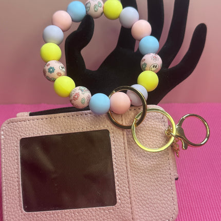 A pink faux-leather card holder wallet with a clear ID window, attached to a pastel beaded bracelet keychain with gold-tone rings and a small keychain charm, displayed on a black hand-shaped stand against a pink background.