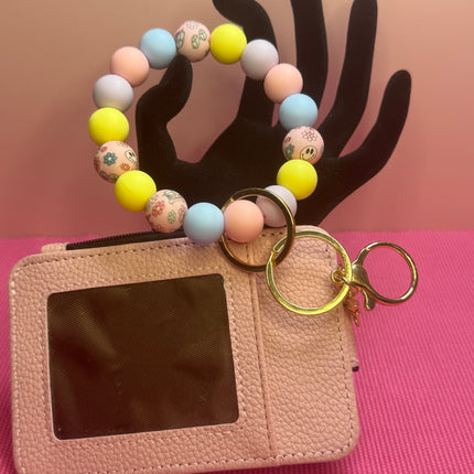 A pink textured leather card holder wallet with a clear ID window, attached to a pastel beaded bracelet keychain with gold key rings, displayed on a black hand-shaped stand against a pink and peach background.