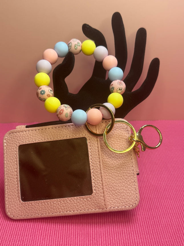 A pink textured leather card holder wallet with a clear ID window, attached to a pastel beaded bracelet keychain with gold key rings, displayed on a black hand-shaped stand against a pink and peach background.