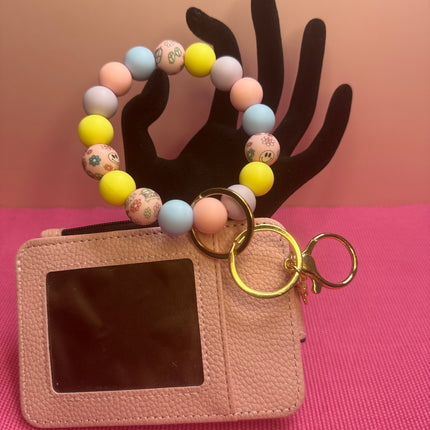 A pink faux-leather card holder wallet with a clear ID window, attached to a pastel beaded wrist strap and gold key rings, displayed on a pink background with a black hand-shaped prop.