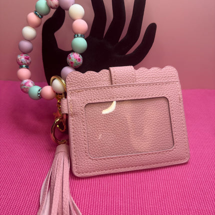 A pink textured faux-leather ID badge holder with a scalloped top edge, a clear plastic window, a matching pink tassel, and a beaded wrist strap in pastel pink, mint green, lavender, and white. The product is displayed on a pink textured surface with a black hand-shaped prop against a pink background.