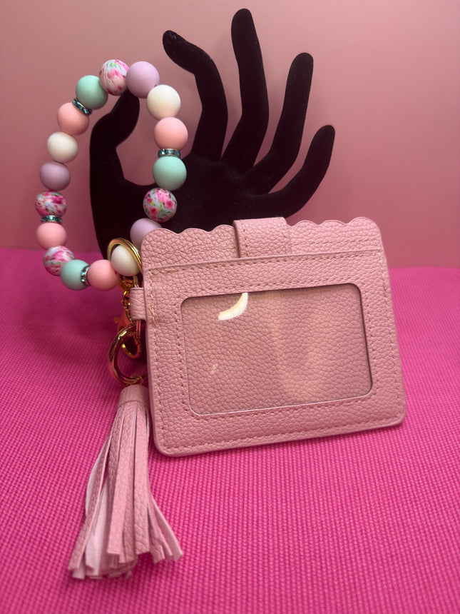 A pink textured faux-leather ID badge holder with a scalloped top edge, a clear plastic window, a matching pink tassel, and a beaded wrist strap in pastel pink, mint green, lavender, and white. The product is displayed on a pink textured surface with a black hand-shaped prop against a pink background.