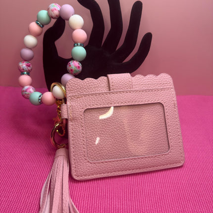 A pink textured faux-leather ID badge holder with a clear window, shown with a matching pink tassel and a beaded wrist strap in pastel colors, displayed on a black hand-shaped stand against a pink background.
