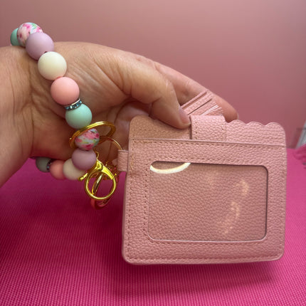 A hand holds a small pink textured card holder with a clear ID window and a beaded wrist strap with gold key rings. The background is a solid pink surface and wall.