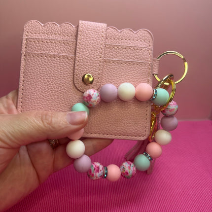 A hand holds a light pink faux-leather card holder wallet with scalloped edges, a snap button, card slots, and a matching beaded wrist strap with pastel pink, lavender, mint, and white beads, attached to gold-tone hardware, against a pink background.