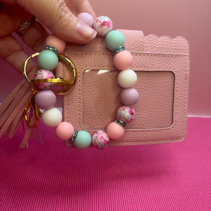 A hand holds a pastel beaded keychain with a tassel and gold keyring in front of a pink textured card holder. The beads are light pink, mint green, lavender, and white marbled.