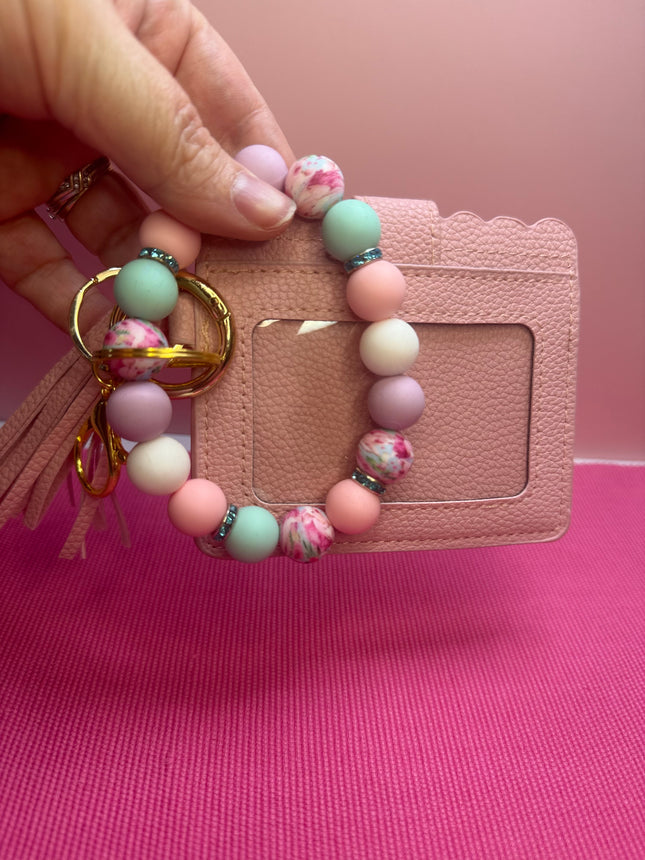 A hand holds a pastel beaded keychain with a tassel and gold keyring in front of a pink textured card holder. The beads are light pink, mint green, lavender, and white marbled.