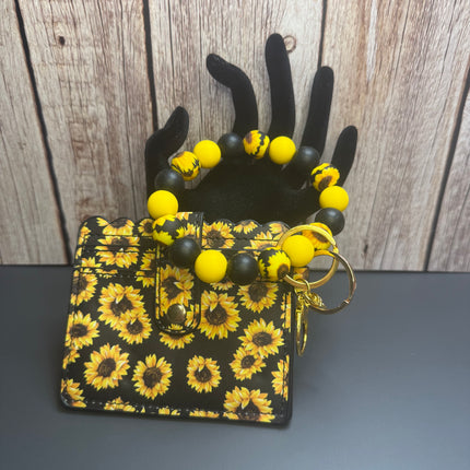 A small black wallet with a yellow sunflower print and a matching beaded bracelet with yellow and black beads, displayed on a black hand-shaped stand against a rustic wood background.