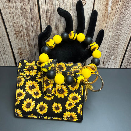 A black & yellow sunflower wallet and matching beaded wristlet  keychain set with yellow and black sunflower patterns, displayed on a black hand-shaped stand against a split wood and gray background.