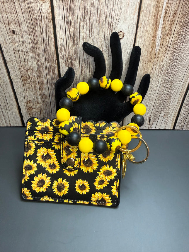 A black & yellow sunflower wallet and matching beaded wristlet  keychain set with yellow and black sunflower patterns, displayed on a black hand-shaped stand against a split wood and gray background.