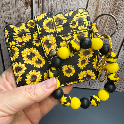 Sunflower faux leather wristlet wallet combo featuring a black sunflower print wallet, clear ID holder, silicone beaded wristlet, gold hardware, and matching sunflower accent beads.