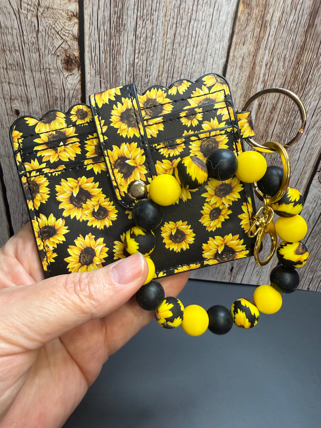 Sunflower faux leather wristlet wallet combo featuring a black sunflower print wallet, clear ID holder, silicone beaded wristlet, gold hardware, and matching sunflower accent beads.