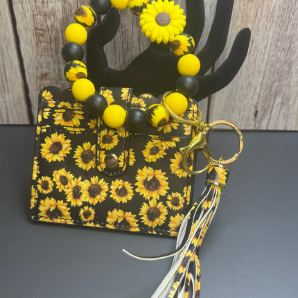 A small black and yellow sunflower-patterned handbag with a matching beaded handle and tassel keychain, displayed on a black hand-shaped stand against a wood plank background.