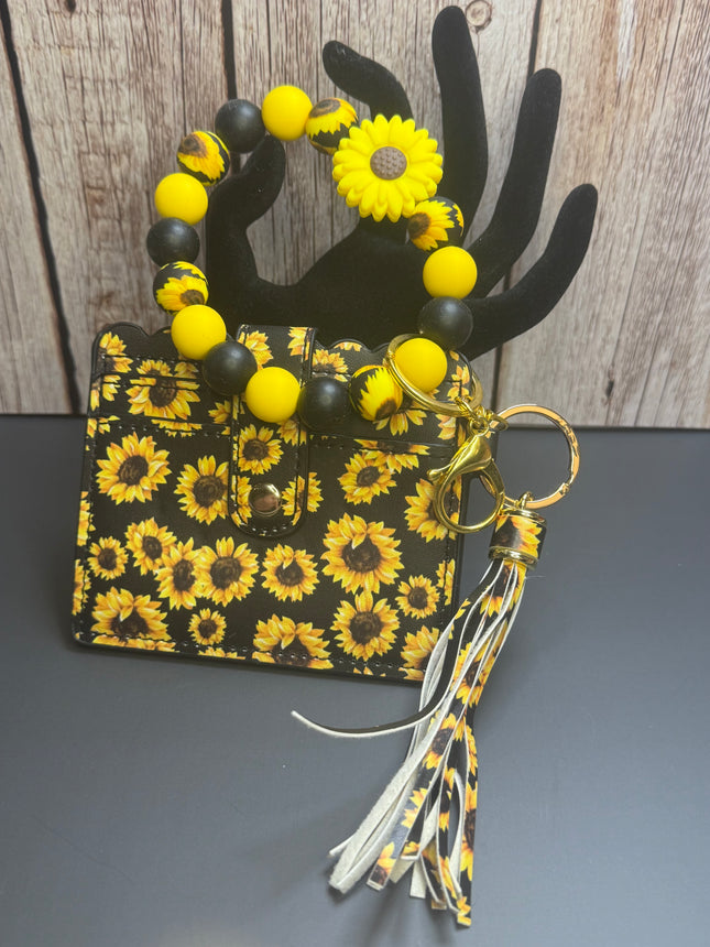 A small black and yellow sunflower-patterned handbag with a matching beaded handle and tassel keychain, displayed on a black hand-shaped stand against a wood plank background.
