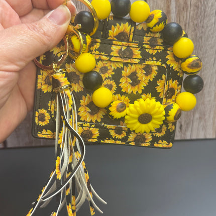 A hand holds a small card holder wallet featuring a yellow and black sunflower pattern, decorated with a matching beaded bracelet, gold key rings, and a tassel with white, black, and yellow strands.