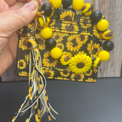 A hand holds a sunflower-themed keychain holder with a matching card holder. The holder features a black background with a yellow and brown sunflower pattern, decorated with yellow and black beads, a yellow flower charm, and a white and yellow tassel.