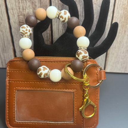 A brown faux-leather card holder wallet with a beaded wrist strap keychain. The wallet has a clear ID window and a gold key ring with a lobster clasp.
