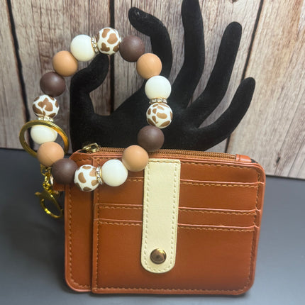 Handmade caramel faux leather wallet with cow print silicone beaded wristlet featuring a clear ID window, zippered coin pocket, card slots, gold hardware, and western-inspired neutral beads.