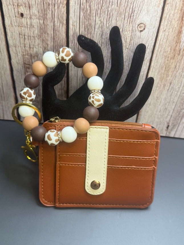 Handmade caramel faux leather wallet with cow print silicone beaded wristlet featuring a clear ID window, zippered coin pocket, card slots, gold hardware, and western-inspired neutral beads.