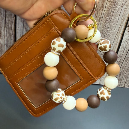 A hand holds a brown leather card holder wallet next to a matching beaded keychain. The keychain features matte brown, cream, and tan round beads, some with a giraffe print pattern, and gold-tone metal rings.