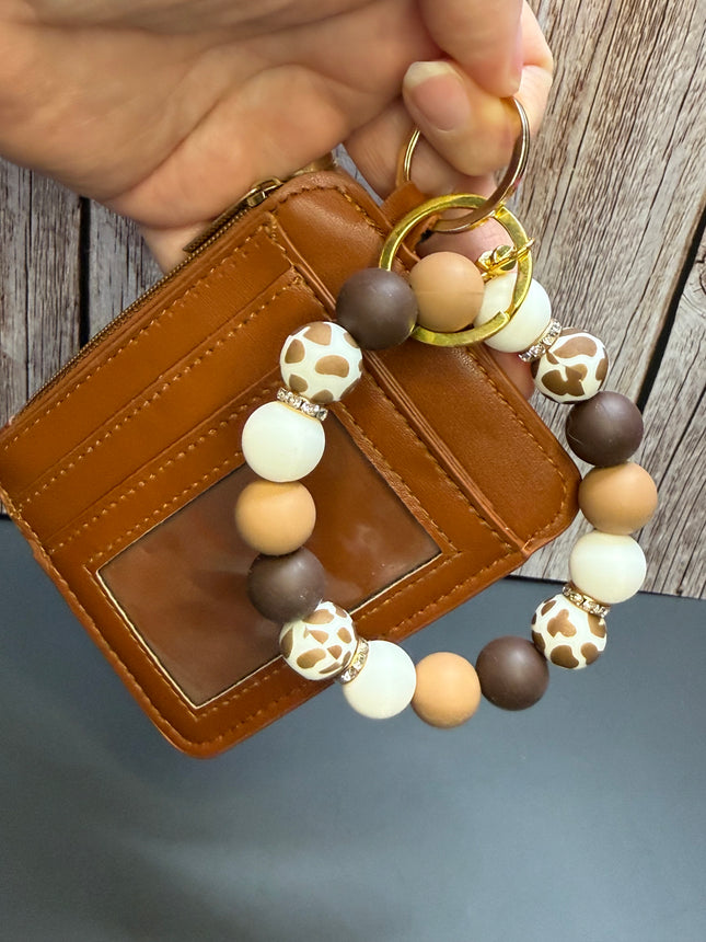 A hand holds a brown leather card holder wallet next to a matching beaded keychain. The keychain features matte brown, cream, and tan round beads, some with a giraffe print pattern, and gold-tone metal rings.