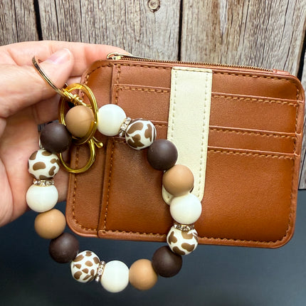 A hand holds a brown faux-leather card holder wallet with a matching beaded keychain charm. The wallet has a vertical cream stripe and card slots, while the keychain features brown, cream, and spotted pattern beads on a gold ring.