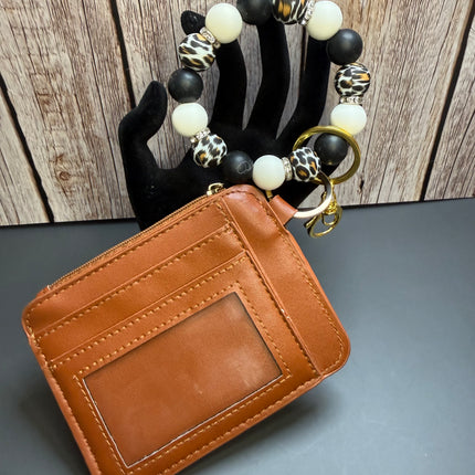 A brown leather card holder wallet with an ID window, attached to a beaded bracelet with black, white, and leopard print beads, displayed on a black hand form against a wood panel background.