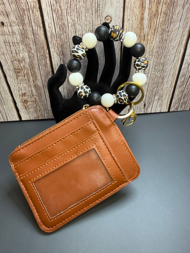 A brown leather card holder wallet with an ID window, attached to a beaded bracelet with black, white, and leopard print beads, displayed on a black hand form against a wood panel background.