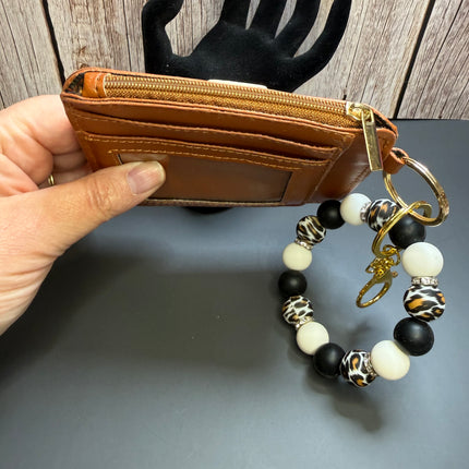 A tan leather zippered card holder wallet with a gold-tone key ring and a black, white, and leopard-print beaded keychain, held by a hand in front of a black hand-shaped display and wood panel background.
