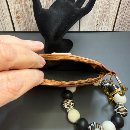 A close-up of a hand holding open a small brown leather zip pouch, with a beaded bracelet draped from the zipper. The bracelet features black, white, and leopard-print beads, and a gold-tone clasp.