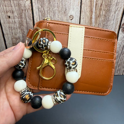 A hand holds a tan faux-leather card holder wallet with a matching beaded keychain charm. The wallet has a vertical cream stripe and card slots, while the keychain features black, white, leopard print, and metallic gold beads with a gold key ring and clasp.